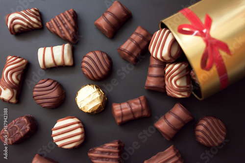 Cioccolatini in diverse forme e colori versato dalla scatola rotonda d'oro con un fiocco rosso su sfondo nero. Tra tutti i dolci hanno un oro avvolto.  photo