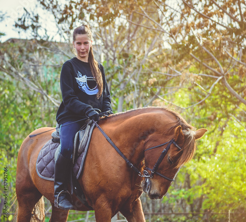 Young beautiful brunette girl rides a horse on a warm and sunny autumn day. Portrait of a pretty young woman on the horse, wearing tall boots and gloves.