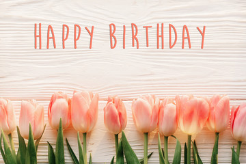 happy birthday text sign on pink tulips on white rustic wooden background flat lay. spring top view of flowers in soft morning light with space for text. greeting card concept