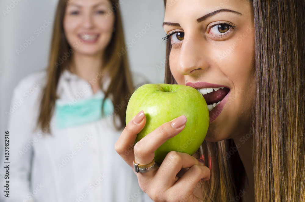 Pretty young brunette with perfect white teeth biting apple, dentist in background. Pretty young brunette with perfect white teeth biting apple, dentist in background.