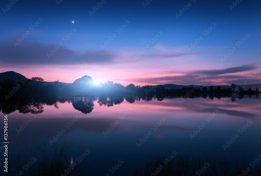 Mountain lake with moonrise at night. Night landscape with river, trees, hills, moon and colorful blue sky with clouds reflected in water in dusk. Beautiful nature background in twilight. Travel