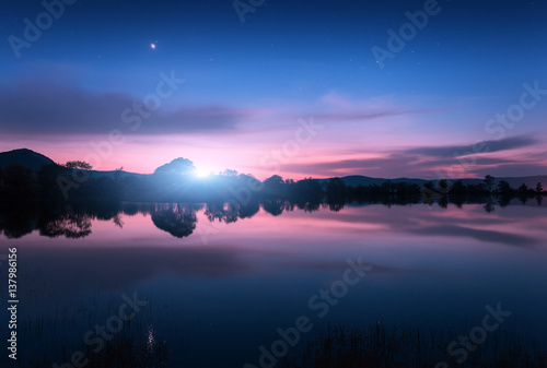 Fototapeta Naklejka Na Ścianę i Meble -  Mountain lake with moonrise at night. Night landscape with river, trees, hills, moon and colorful blue sky with clouds reflected in water in dusk. Beautiful nature background in twilight. Travel