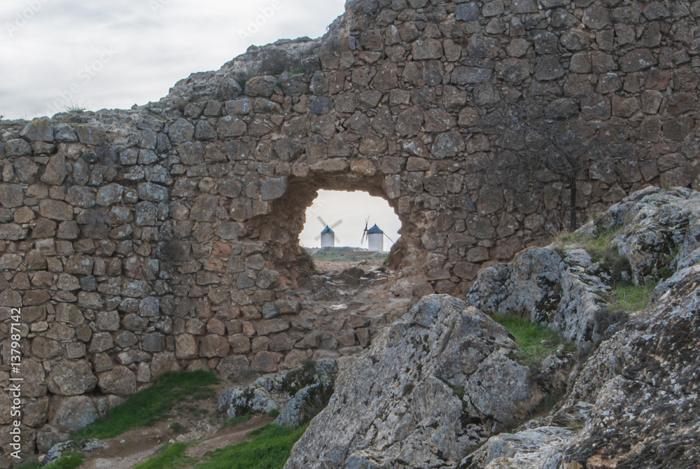 White old windmills on the hill near Consuegra (Castilla La Mancha, Spain), a symbol of region and journeys of Don Quixote (Alonso Quijano) and a town on cloudy day, a view through a hole in the wall.