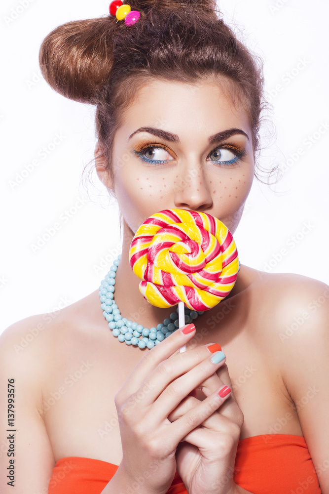 Beautiful brunette sweet woman with colorful make up and nail polish, and cute bun hairstyle. Eating lollipop. white background