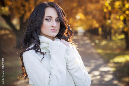 Beautiful caucasian brunette young woman in warm autumn day at park. Yellow leaves and sunshine. Fall season. Woman in white sweater, smiling. Copy space