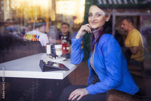 Brunette woman in business clothes  grey sweater and blue jacket sitting at the cafe near the window in european city drinking latte coffee and working using her tablet and smartphone. copy space