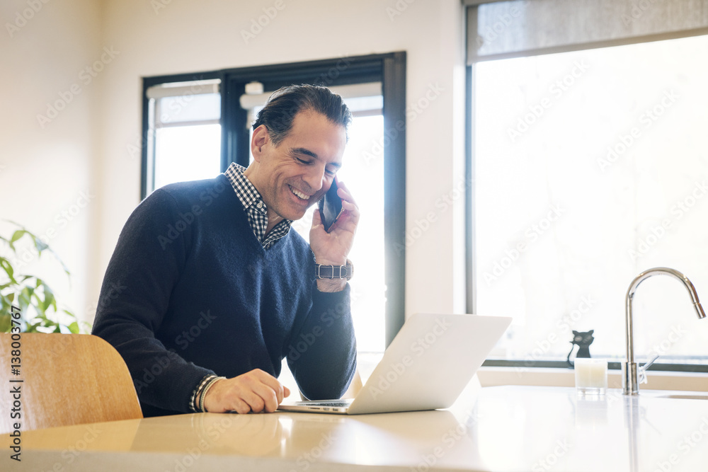 Happy man talking on mobile phone while sitting by laptop at table Happy man talking on mobile phone while sitting by laptop at table