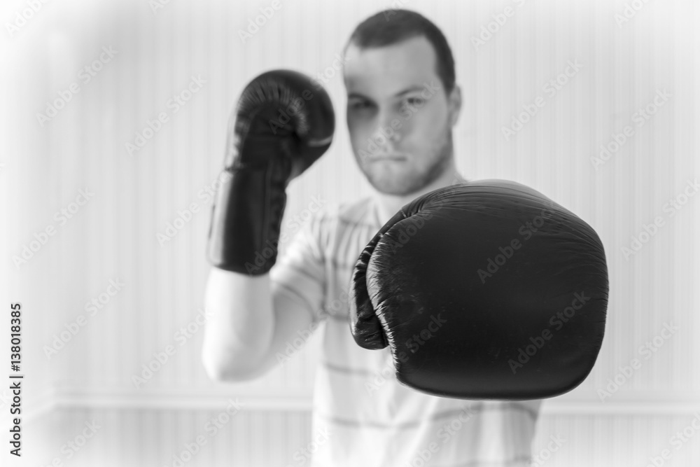 horizontal black and white  close up image of a young caucasian adult male wearing boxing gloves in a fighting stance and throwing a punch towards camera.