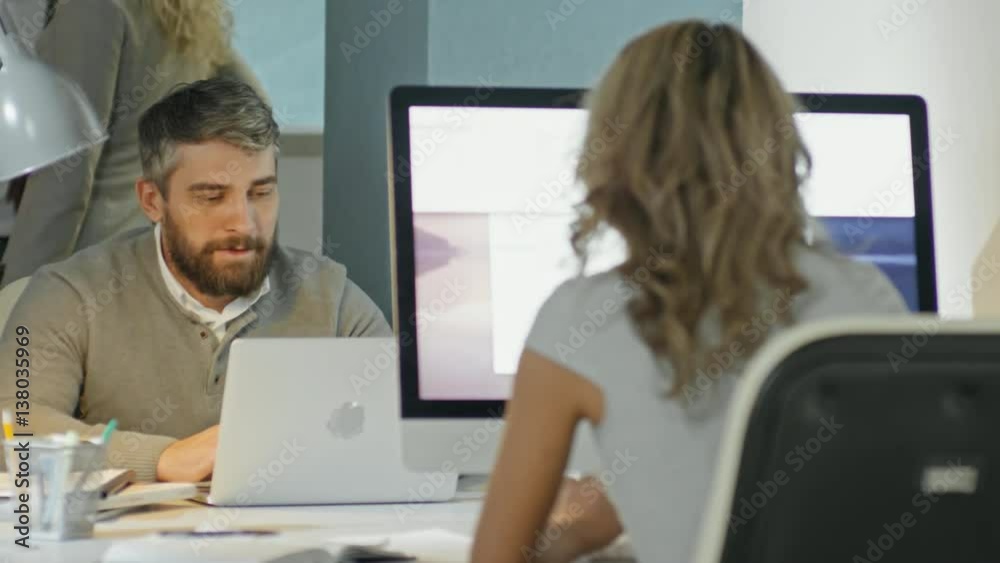 PAN of businessman with beard working on laptop and discussing work with female colleague sitting opposite him PAN of businessman with beard working on laptop and discussing work with female colleague sitting opposite him