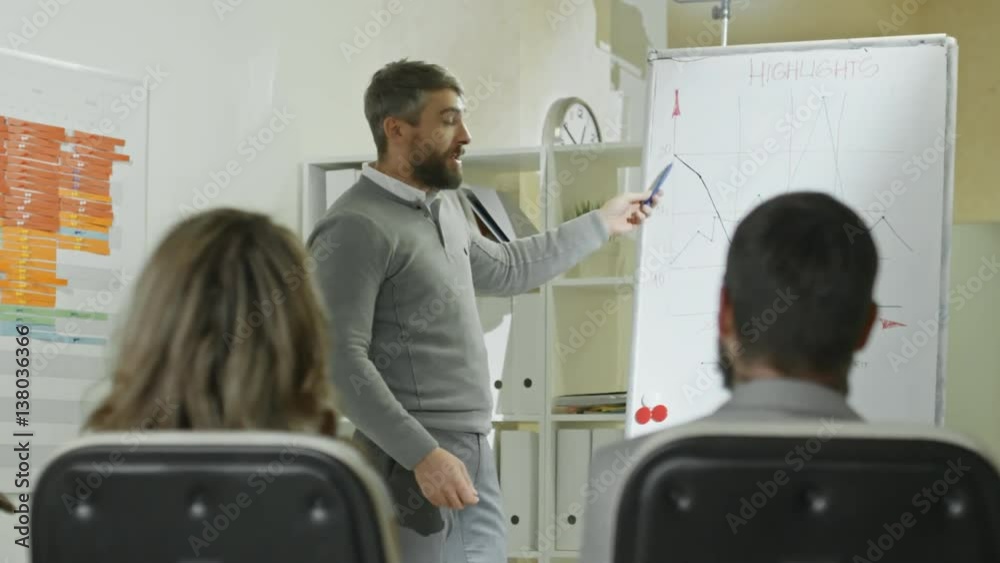 Handheld shot of bearded businessman gesturing and speaking about company financial growth during presentation before audience 
