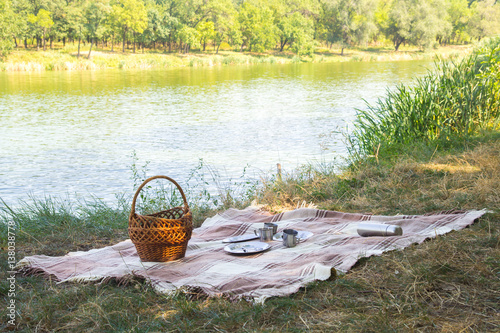 picnic set, metal Cutlery, thermos, plates tea cups. brown plaid and napkin from the lake in the background. green grass. Sunny summer