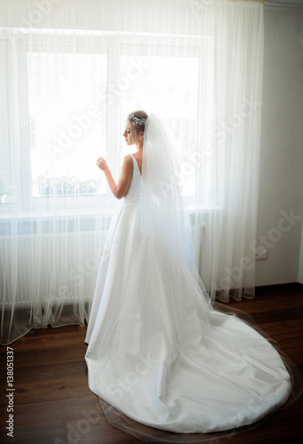 Bride in long white dress with long veil stands before bright window on wooden floor