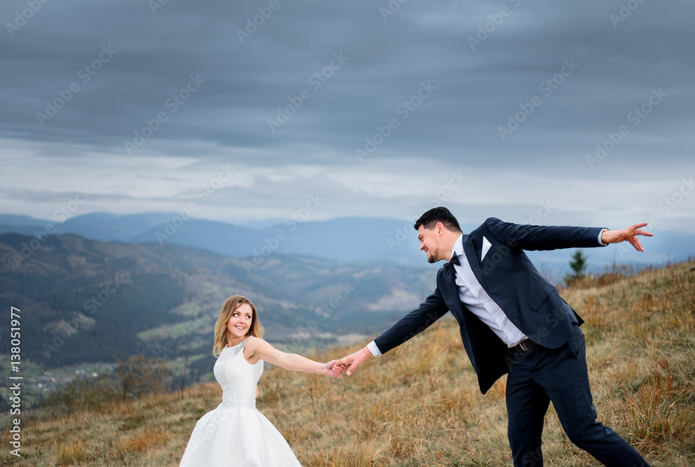 Bride holds groom's hand while they walk down the hill in a cloudy day