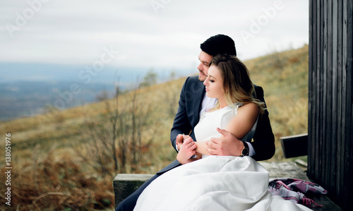 Groom holds bride tender while they rest on old wooden bench on the hill
