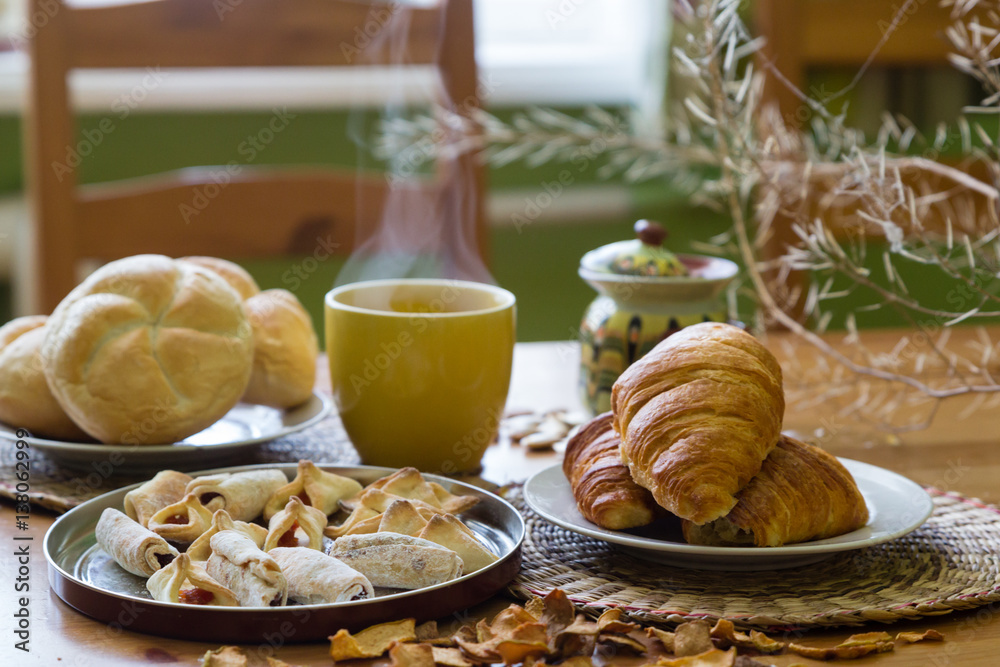 Black tea in yellow mug with croissants, buns and homemade cookies on wooden table