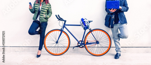 Couple of commuters with bike holding mobile phone and pc notebook leaning on white urban wall - Concept of everyday life going at  work or study place in the city and the modern technology usage