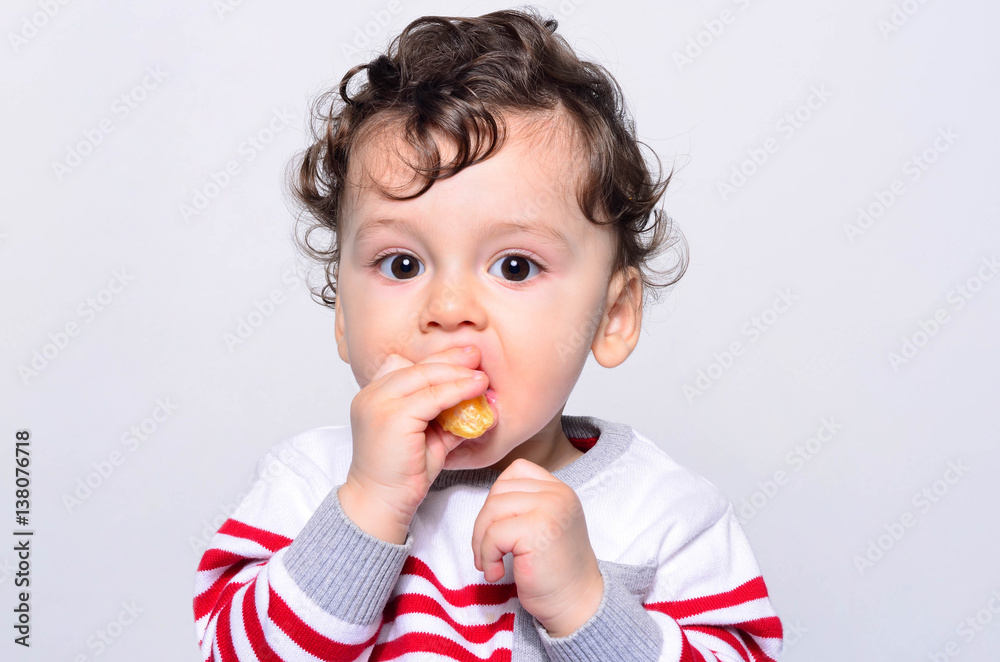 Portrait of a cute baby eating orange. One year old kid eating fruits by himself. Adorable curly hair boy being hungry biting on a slice of orange. Portrait of a cute baby eating orange. One year old kid eating fruits by himself. Adorable curly hair boy being hungry biting on a slice of orange.