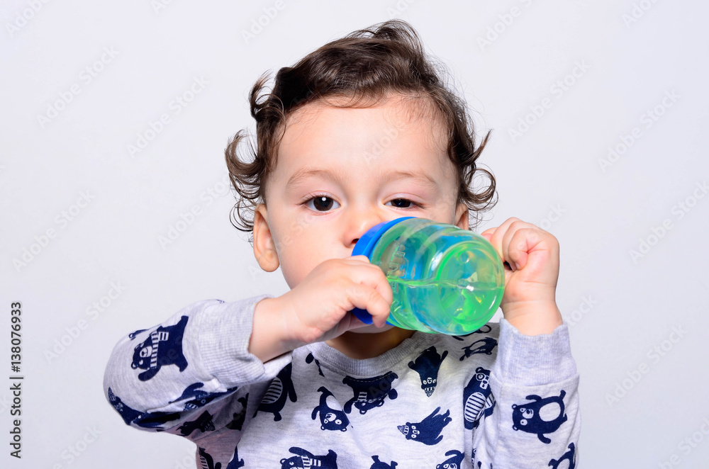 Portrait of a cute toddler drinking water from the bottle. One year old kid holding up the baby cup. Adorable curly hair boy being thirsty. Portrait of a cute toddler drinking water from the bottle. One year old kid holding up the baby cup. Adorable curly hair boy being thirsty.