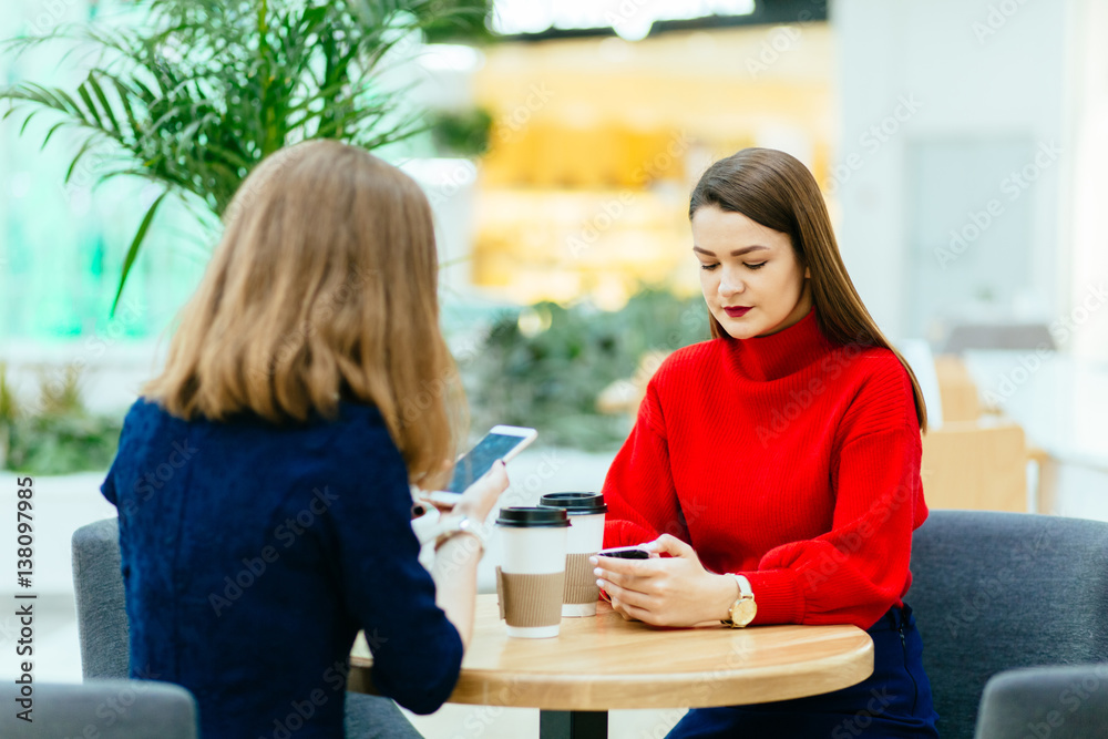Two friends using phone and having a coffee in shopping center. Social concept about people. Two friends using phone and having a coffee in shopping center. Social concept about people.