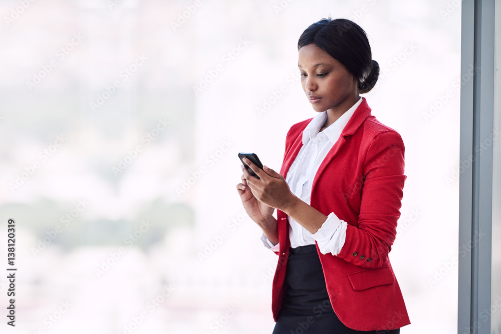 Young formally dressed adult woman busy using her mobile phone while standing in front of large glass windows while wearing a bright red blazer with copy space to her left. Young formally dressed adult woman busy using her mobile phone while standing in front of large glass windows while wearing a bright red blazer with copy space to her left.