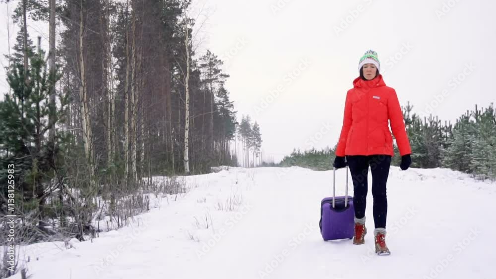 young woman in bright red jacket and knitted hat leave home with packed staff in suitcase and go away along snowy road during snowstrom in winter with very sad and angry emotions on face
