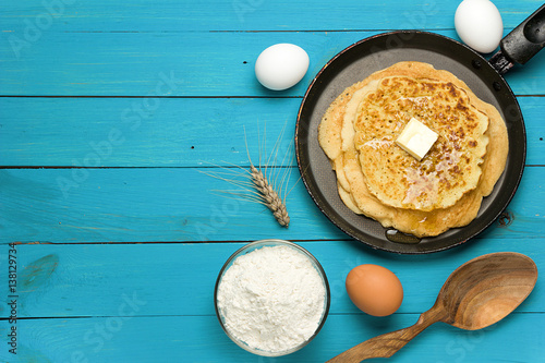 Hot delicious pancakes in frying pan on blue wooden table with flour and eggs