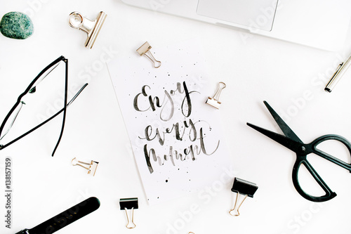 White office desk workspace with quote Enjoy Every Moment and office supplies. Laptop, scissors, pen, clips, glasses on white background. Flat lay, top view.