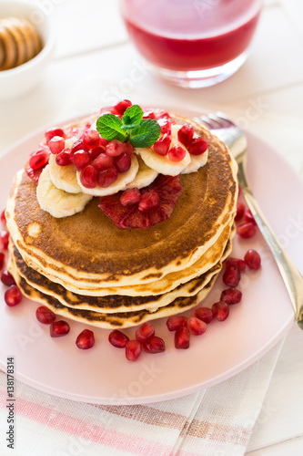 Traditional breakfast: stack of pancakes with banana slices and garnet seeds on white wooden table. Selective focus
