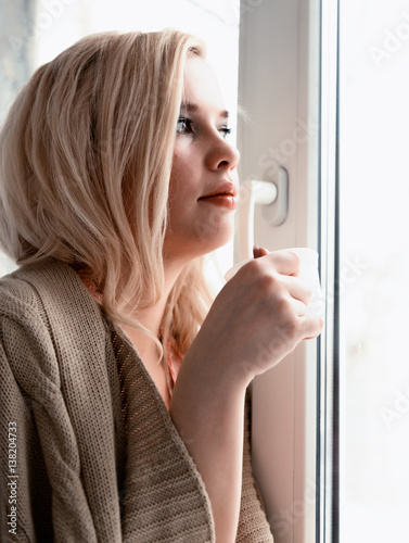 young blonde woman is dreaming about something while sitting on the window-sill. she holding a tea cup and drinking