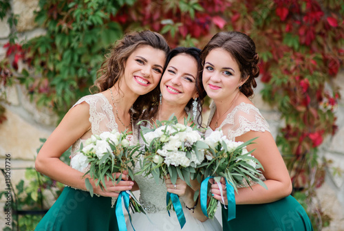Bridesmaids in green skirts stand behind pretty bride before wall with red ivy