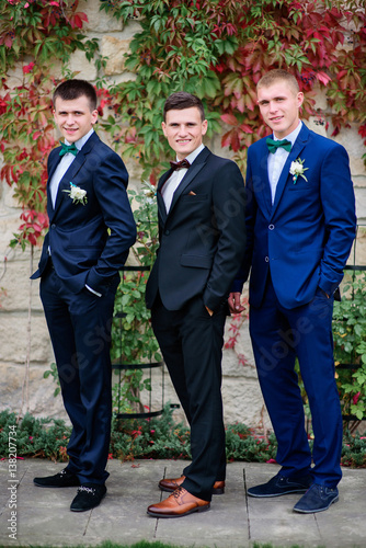 Groomsmen in blue suits and groom in black one pose before green wall outside