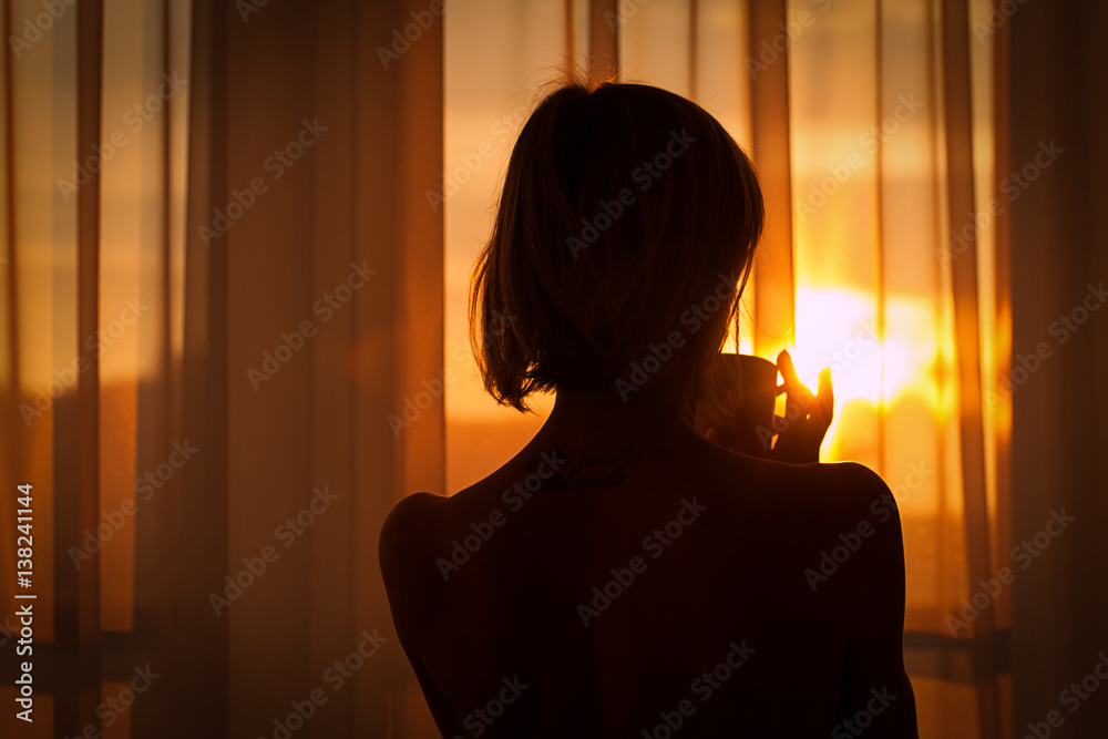 Woman in the morning. Attractive sexy woman with neat body is looking at the sunrise standing near the window in her home and having a perfect cozy morning with a cup of coffee.