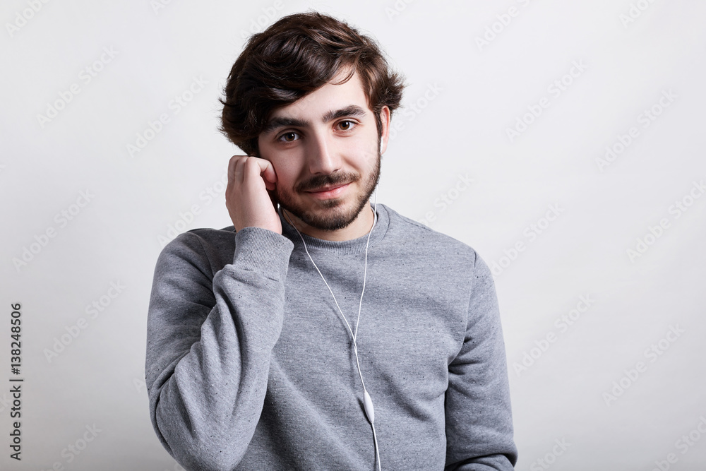 Young handsome bearded hipster with trendy hairstyle standing over white background wearing casual grey sweater enjoying listening to music holding his hand on earphones