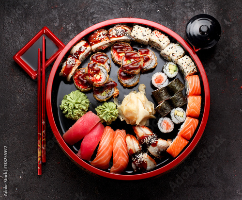 Japanese cuisine. Sushi roll and nigiri set on a round wooden plate with soy sauce and red chopsticks over black concrete background.