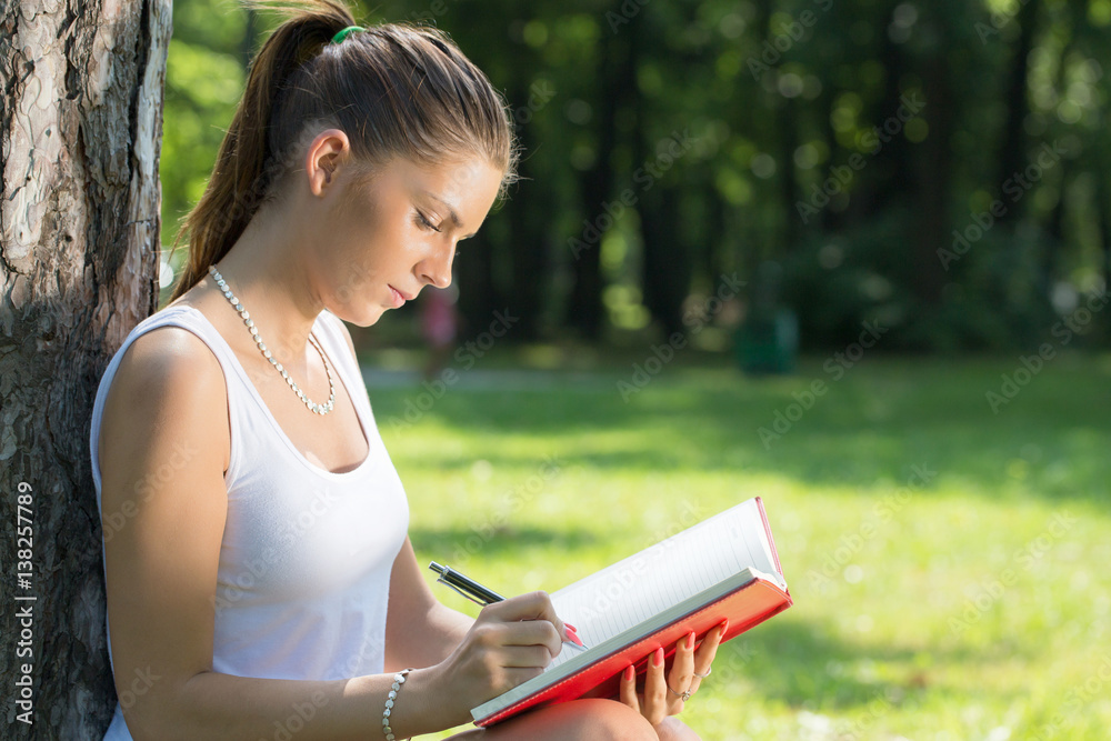 young brunette woman writing to notepad, leaning against a tree young brunette woman writing to notepad, leaning against a tree