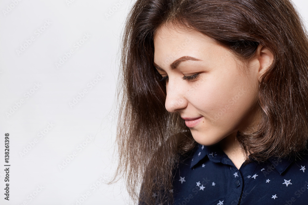 A sideways close-up of beautiful brunette with dark eyebrows, long eyelashes and pure skin dressed in dark shirt looking down with thoughtful expression. People and fashion concept. A sideways close-up of beautiful brunette with dark eyebrows, long eyelashes and pure skin dressed in dark shirt looking down with thoughtful expression. People and fashion concept.