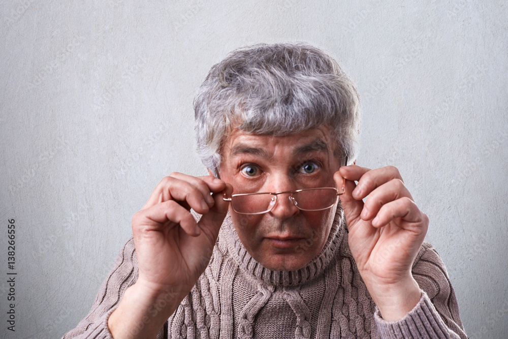 A close-up of surprised adult with gray hair and wrinkles wearing glasses. A senior man touching his eyeglasses looking with wide open eyes into camera being astonished to hear some news A close-up of surprised adult with gray hair and wrinkles wearing glasses. A senior man touching his eyeglasses looking with wide open eyes into camera being astonished to hear some news