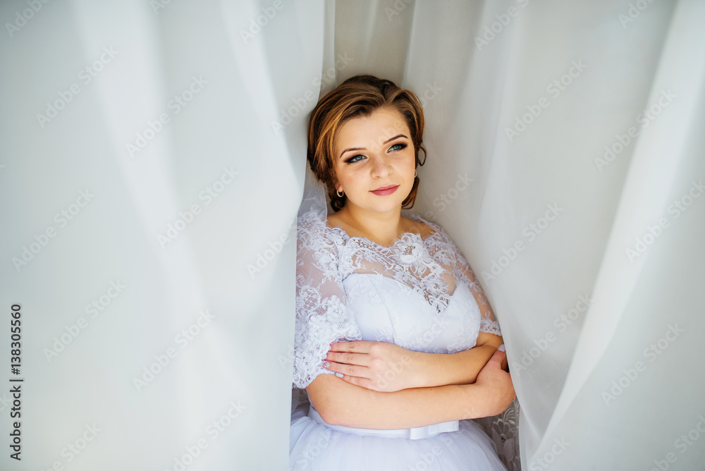 Cute blonde bride posed on curtains at wedding morning day. Cute blonde bride posed on curtains at wedding morning day.