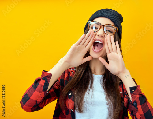 Hipster girl screaming like in megaphone holding hands near her face with open mouth. Young woman touts everyone over yellow background.