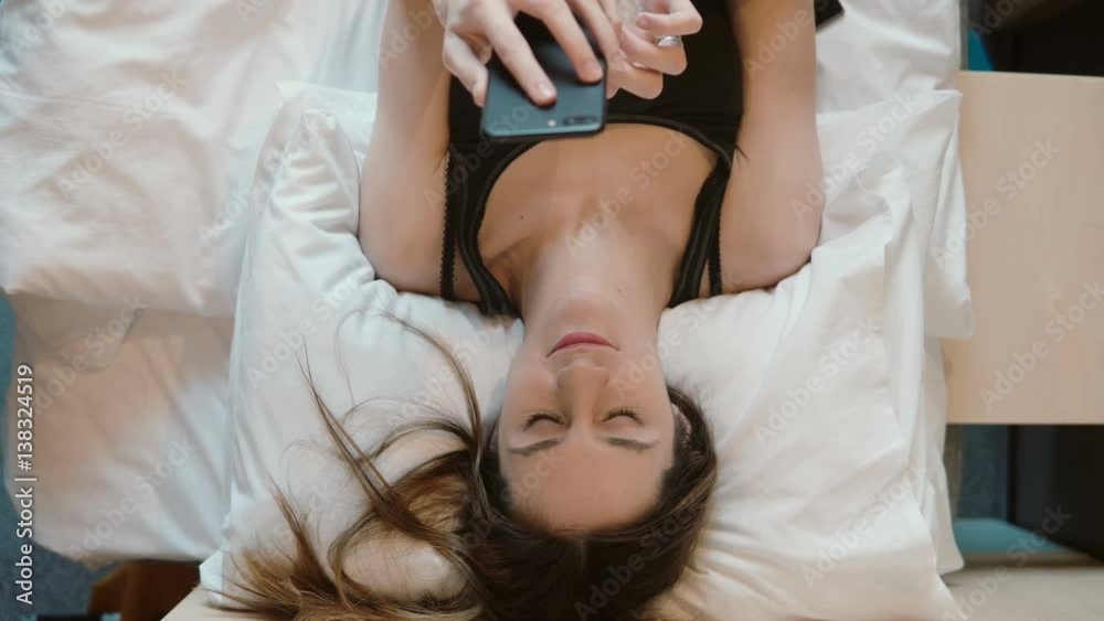 Young woman lying on the bed and holding smarphone. Girl browsing the Internet, chatting with someone. Young woman lying on the bed and holding smarphone. Girl browsing the Internet, chatting with someone.