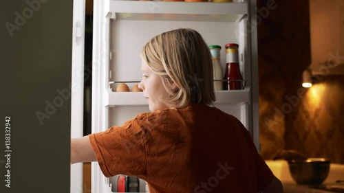 Young blonde Caucasian kid opening fridge biting apple and taking bottle of yogurt in evening