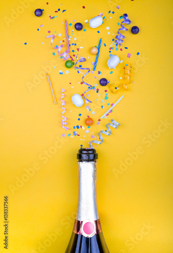 Colorful party attributes fly out from botle of champagne wine. on a yellow background. Flat lie. Celebrate concept. High resolution photo.
