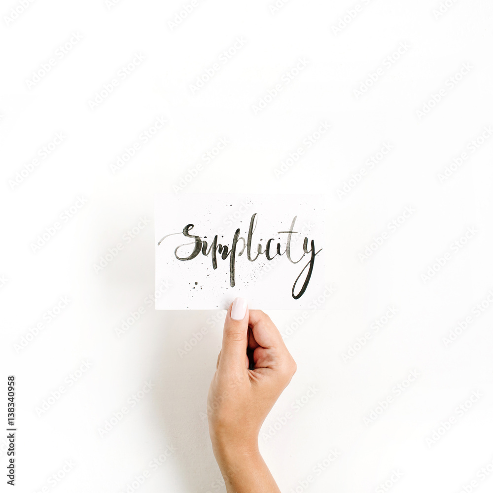 Fototapeta premium Minimal pale composition with girl's hand holding card with word Simplicity written in calligraphic style on paper on white background. Flat lay, top view