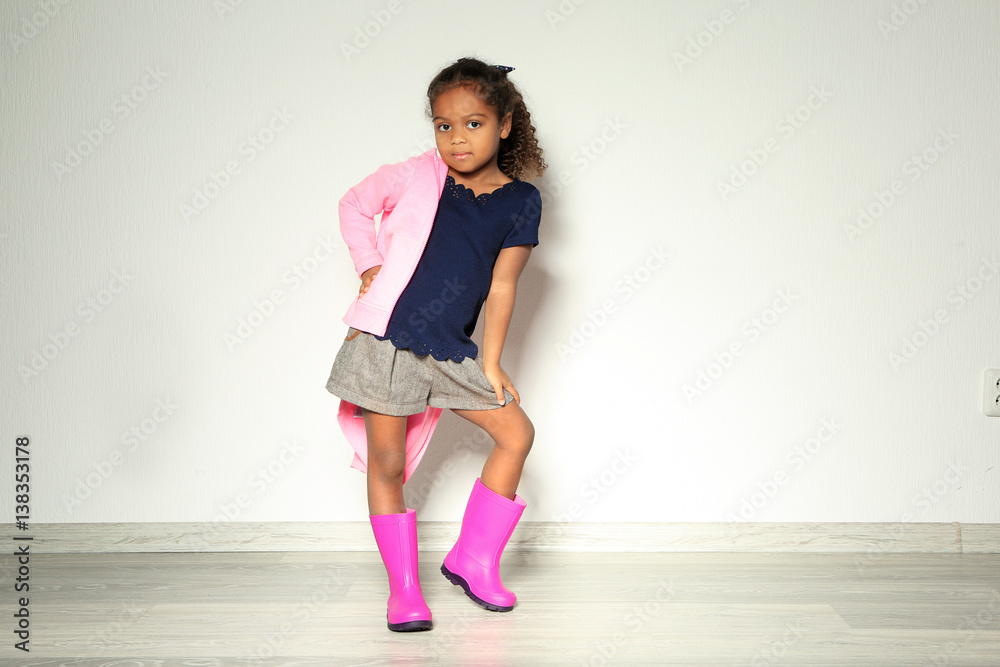 Cute little African American girl in rubber boots against light wall. Fashion concept Cute little African American girl in rubber boots against light wall. Fashion concept