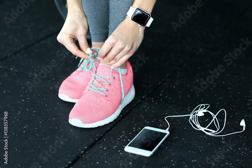 Young woman with fitness tracker and mobile phone tying shoelaces in gym