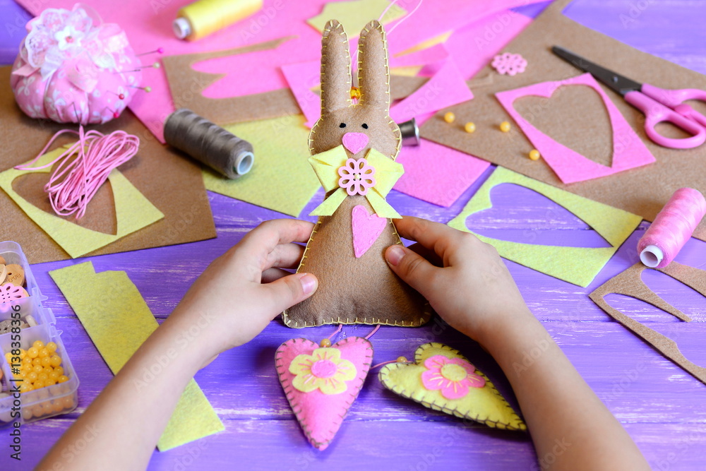 Fototapeta premium Small child holds a felt Easter bunny with hearts in his hands. Child made a cute Easter wall decor made of felt. Easy Easter crafts idea for kids. Craft materials and tools on a wooden table