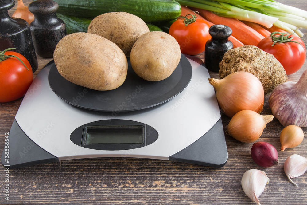 Potatoes on the weighing scales on the table with onions, tomatoes,  cucumbers, garlic, carrots and other vegetables in the kitchen. Healthy  eating and lifestyle. Stock Photo | Adobe Stock