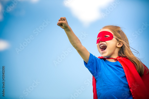 Funny little girl plaing power super hero over blue sky background. Superhero concept.