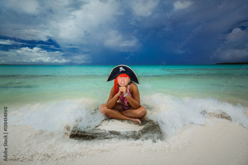 amazing beautiful little pirate girl making funny angry face, sitting on the tropical beach against tranquil ocean and dark dramatic sky background