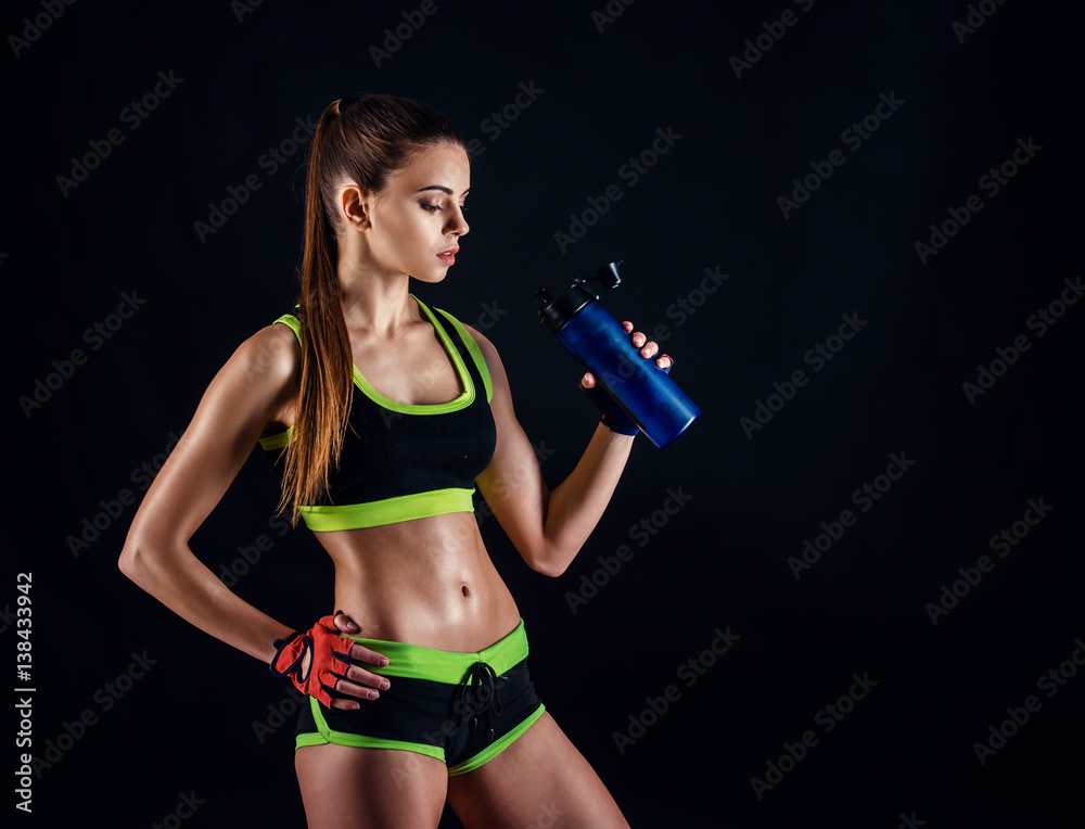Young athletic woman in sportswear with a shaker in studio against black background. Ideal female sports figure. Fitness girl with perfect sculpted muscular and tight body.